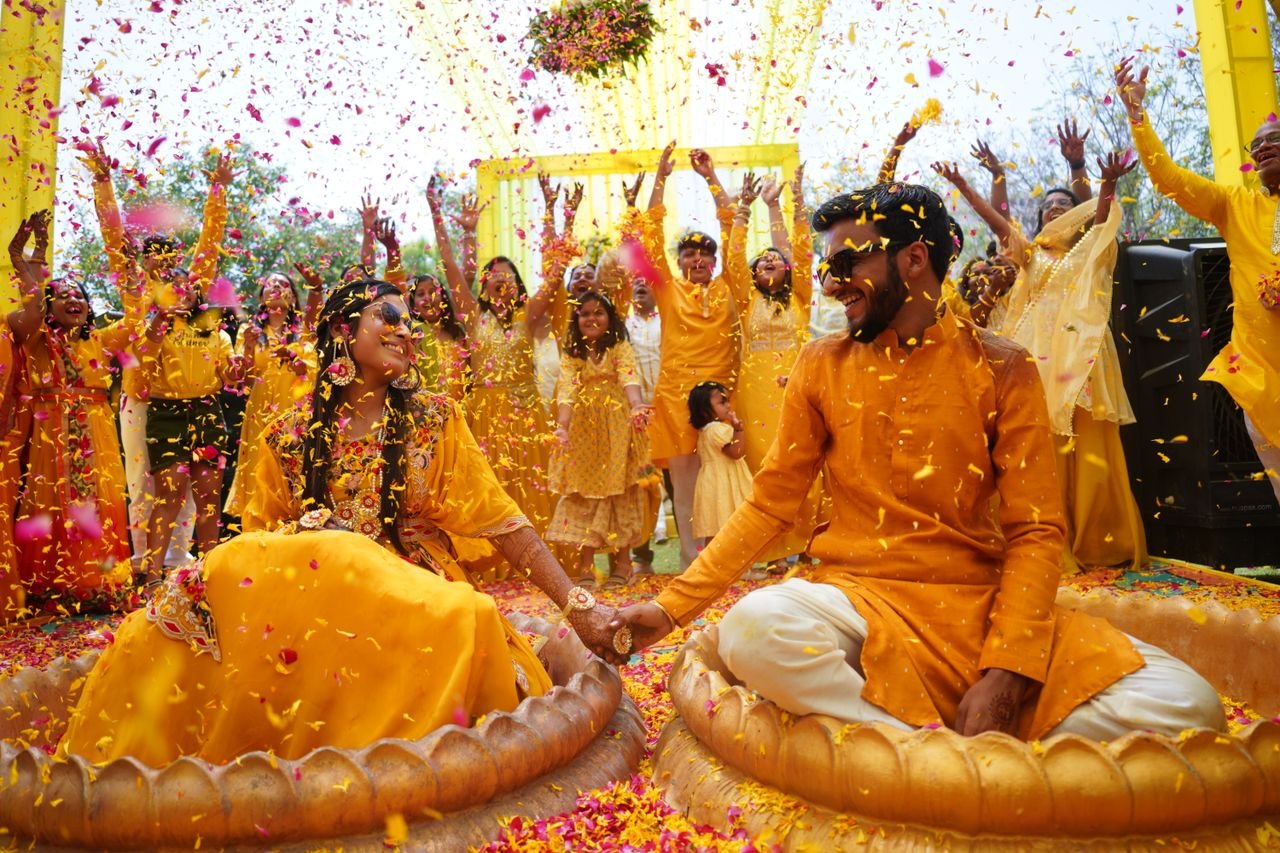 Joyful Indian haldi ceremony with a cheerful couple and guests throwing flower petals outdoors.